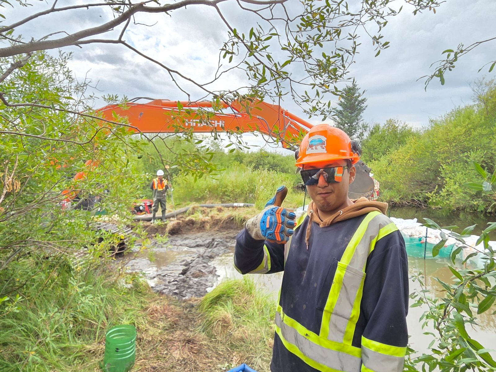 Crew member giving a thumbs up in front of an excavator at a stream work site
