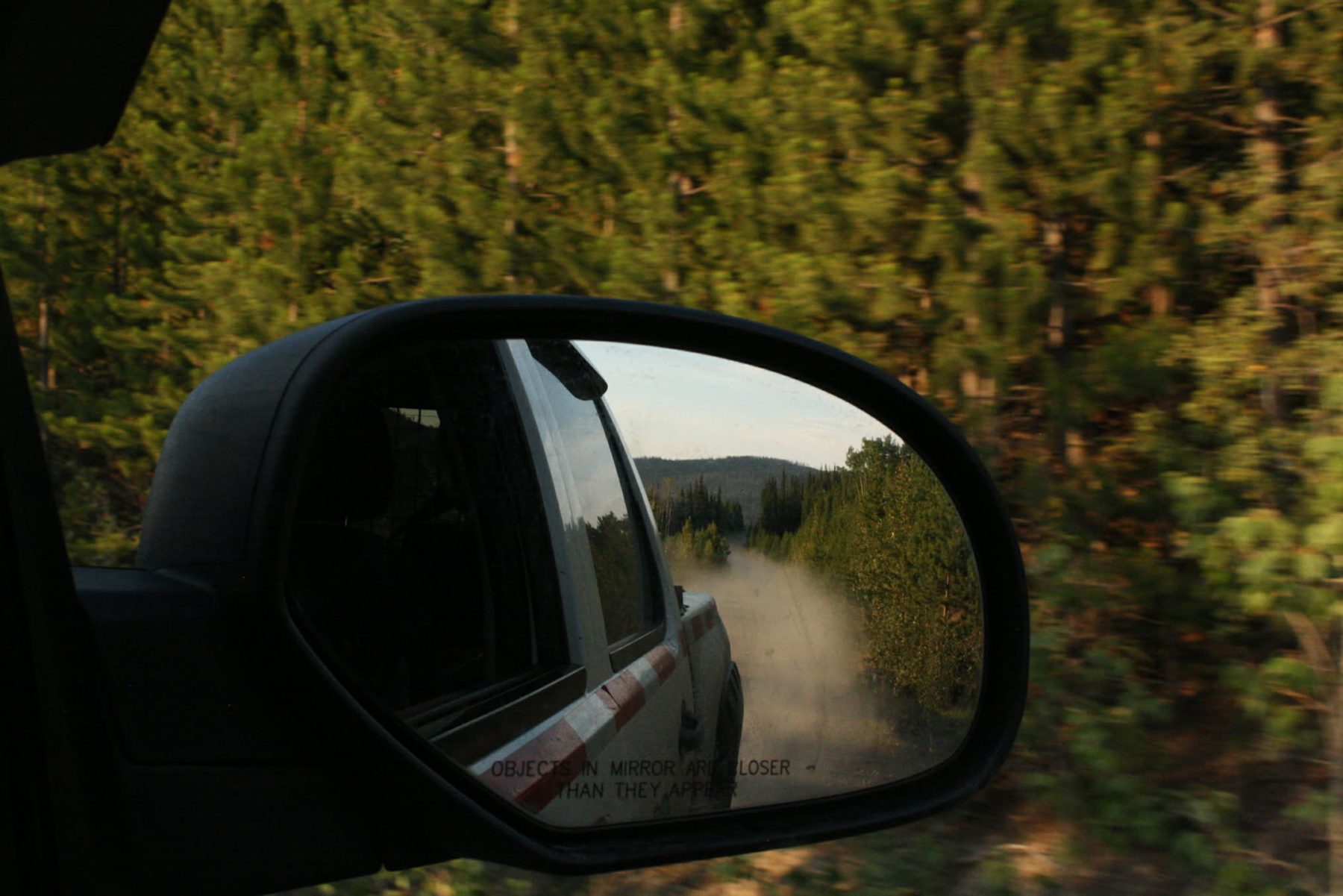 Truck mirror showing a dust cloud on a gravel road through pine