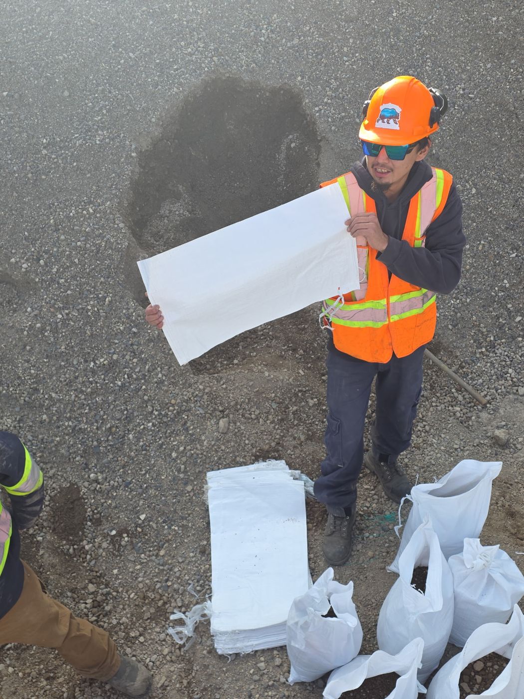 Crew member holding an empty sandbag beside filled bags on gravel